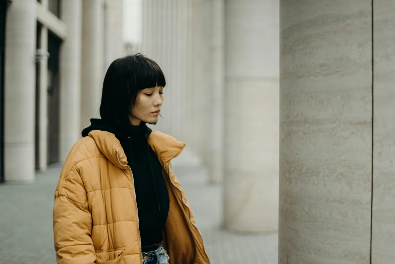 a woman standing in front of a tall building, inspired by Wang E, trending on pexels, postminimalism, puffer jacket, ochre, woman with black hair, ethnicity : japanese