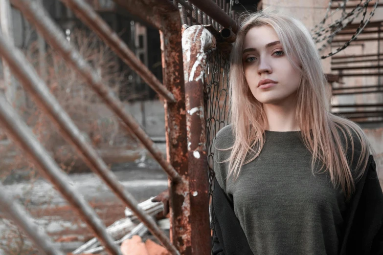 a woman standing in front of a rusted building, a colorized photo, inspired by Elsa Bleda, pexels contest winner, aestheticism, long blonde hair and big eyes, gray skin. grunge, russian girlfriend, an emo girl