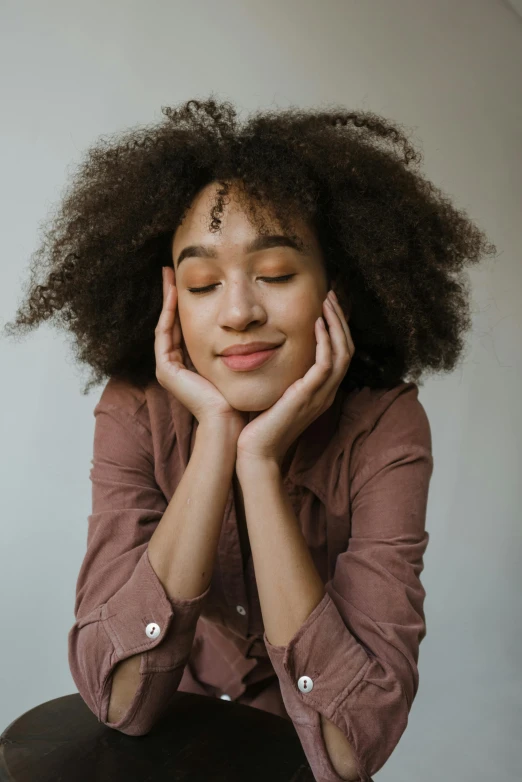 a woman sitting on a stool with her eyes closed, trending on pexels, natural hair, hand on her chin, soft happy smile, with brown skin