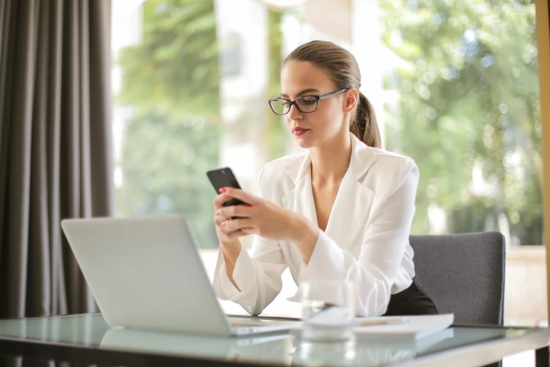 a woman sitting at a table using a cell phone, wearing black rimmed glasses, it specialist, sleek white, morning hour