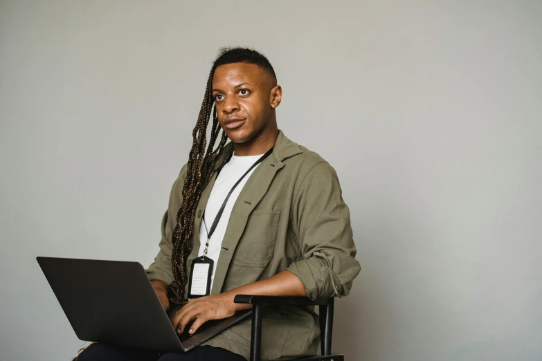 a man sitting in a chair with a laptop, jemal shabazz, on grey background, looking confident, in front of a computer