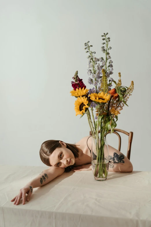 a woman laying on a table next to a vase of flowers, by Winona Nelson, fleshy botanical, kailee mandel, tattooed, trending photo