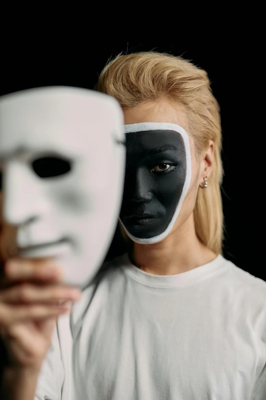 a woman holding a white mask in front of her face, a black and white photo, trending on pexels, antipodeans, half robot and half woman, square, painted face, male and female