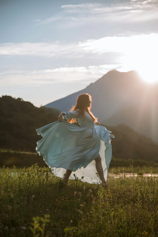 a woman standing on top of a lush green field, unsplash contest winner, romanticism, flowing blue skirt, sun setting, with mountains in background, dancing gracefully