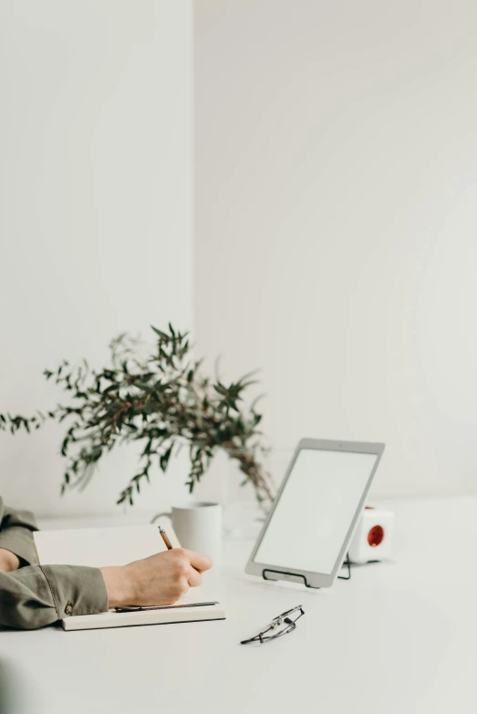 a woman sitting at a table in front of a laptop, white hue, holding a clipboard, inside white room, in a frame