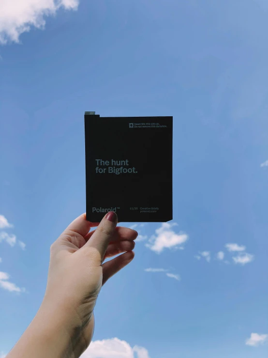 a person holding up a book with a sky background, a polaroid photo, by Maggie Hamilton, pexels contest winner, postminimalism, all black matte product, brochure, diecut, pixelsort