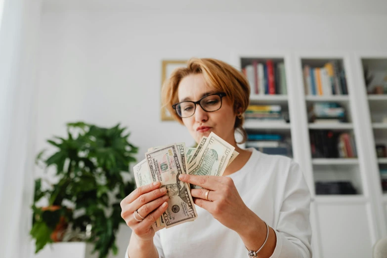 a woman holding a stack of money in front of her face, by Julia Pishtar, pexels contest winner, renaissance, wearing medium - sized glasses, avatar image, at home, fan favorite