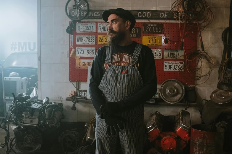 a man with a beard standing in a garage, lowbrow, wearing dirty overalls, official store photo, mechanical accents!