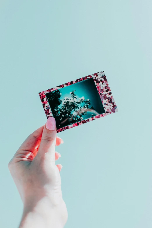 a person holding a picture in their hand, a polaroid photo, by Julia Pishtar, unsplash, serial art, turquoise pink and green, glitter, floral patterned skin, long distance photo