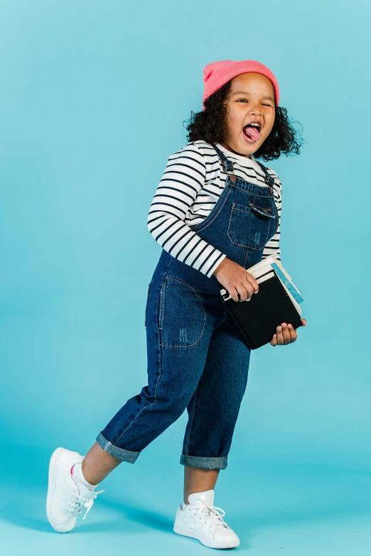 a little girl wearing overalls and a pink hat, an album cover, inspired by Violet Fuller, shutterstock contest winner, happy fashion model, pose(arms up + happy), modest, cyan photographic backdrop