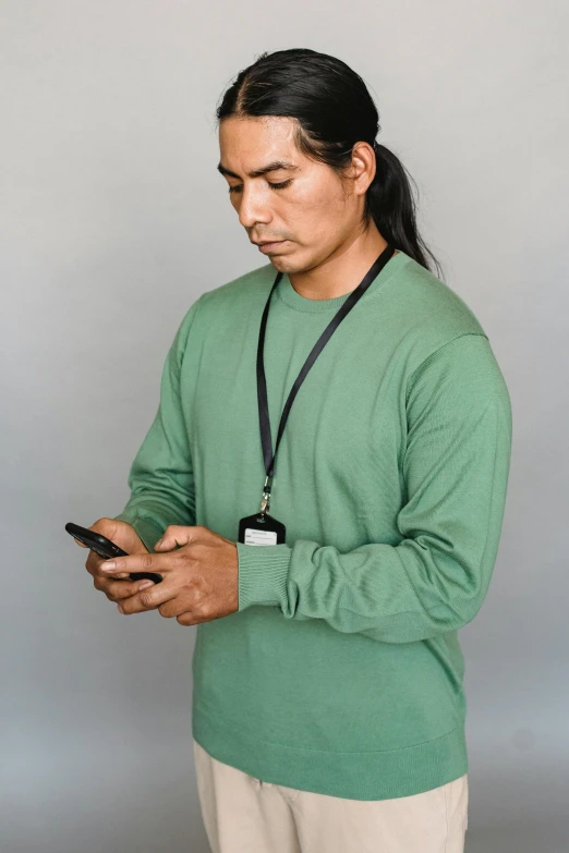 a man in a green shirt looking at his cell phone, a colorized photo, by Gavin Hamilton, wearing a native american choker, on grey background, long sleeves, 2019 trending photo