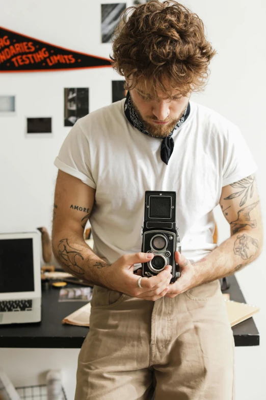 a man standing in front of a desk holding a camera, a tattoo, trending on pexels, photorealism, rolleiflex tlr, ed sheeran, scruffy man, medium format color photography