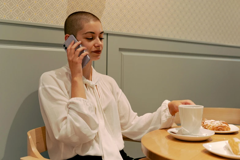 a woman sitting at a table talking on a cell phone, inspired by Clarice Beckett, trending on pexels, antipodeans, brown buzzcut, in style of juergen teller, android coffee shop, female model