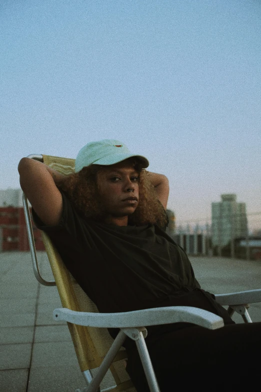 a man sitting in a chair on top of a roof, an album cover, unsplash, renaissance, black teenage girl, wearing a baseball cap backwards, summer evening, headshot