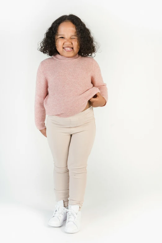 a little girl standing in front of a white background, by Will Ellis, pexels contest winner, wearing casual sweater, widest hips, light blush, wearing pants