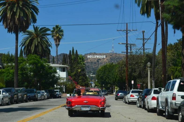 a red car driving down a street next to palm trees, a photo, pexels contest winner, renaissance, hollywood scene, hills in the background, bl, a wooden