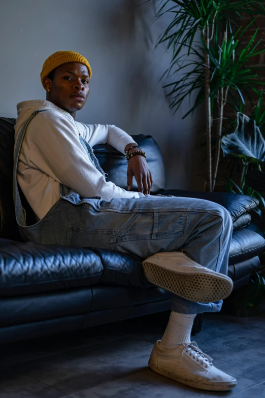 a man sitting on top of a black leather couch, by William Berra, trending on pexels, tyler the creator, non binary model, confident relaxed pose, wearing overalls