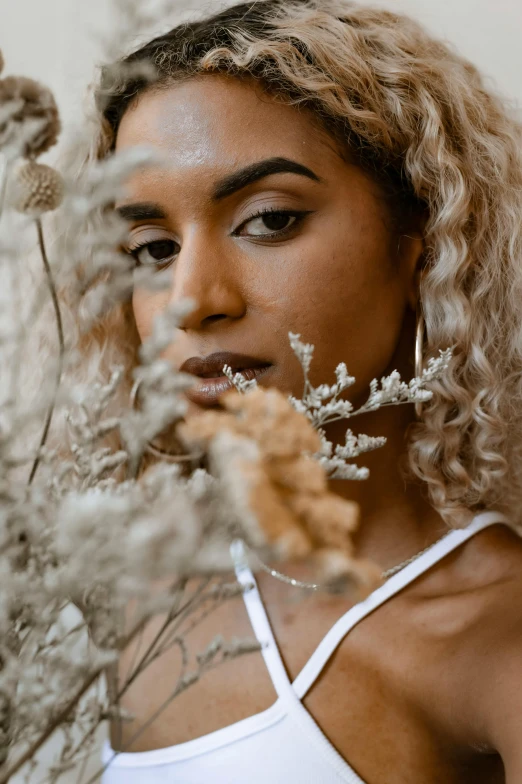 a woman standing in front of a bunch of flowers, inspired by Elsa Bleda, trending on pexels, renaissance, brown skin. light makeup, curly blond, dried plants, close up portrait of woman