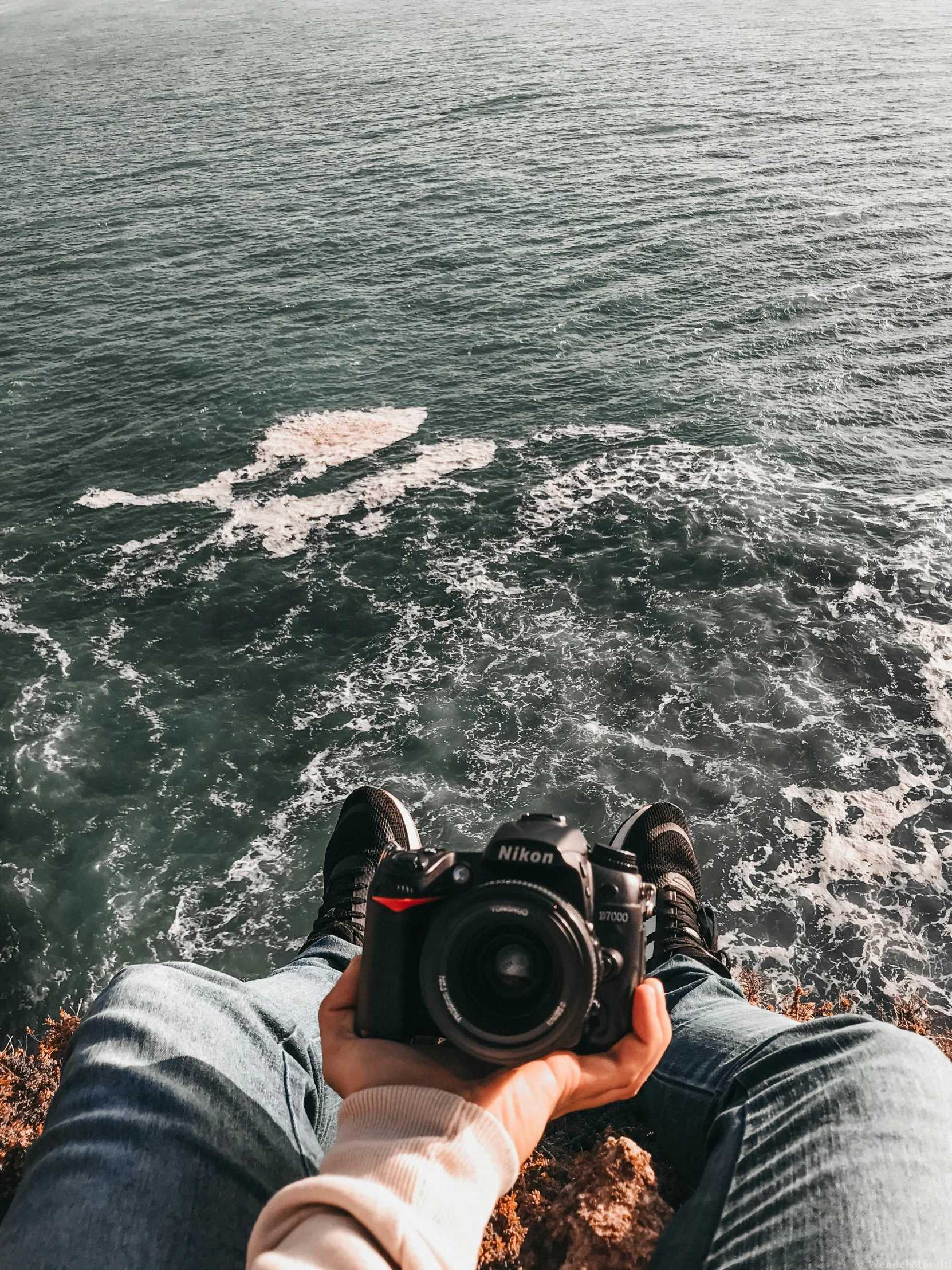 a person taking a picture of the ocean with a camera, by Niko Henrichon, pexels contest winner, sitting down, top down camera angle, sneaker photo, nazare (portugal)