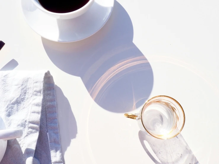 a cup of coffee sitting on top of a white table, by Carey Morris, trending on unsplash, minimalism, sun flare, gold refractions off water, white tablecloth, playful composition