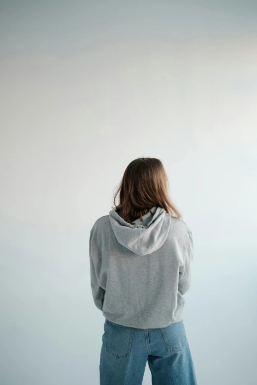 a woman standing in front of a white wall, trending on pexels, wearing a grey hooded sweatshirt, with his back turned, soft hair, loneliness