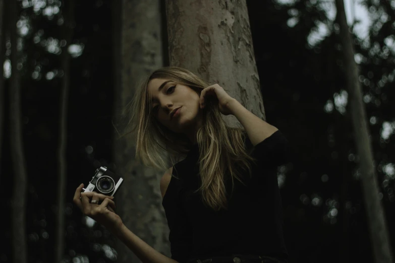 a woman standing next to a tree holding a camera, woman in a dark forest, photoshoot for skincare brand, vintage camera, slightly minimal