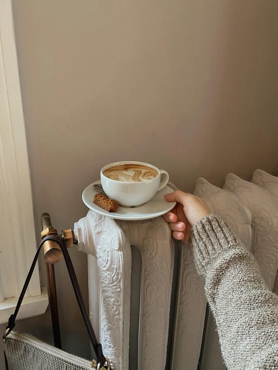 a person holding a cup of coffee near a radiator, by Lucia Peka, trending on instagram, low quality photo, cappuccino, cottagecore, today\'s featured photograph 4k