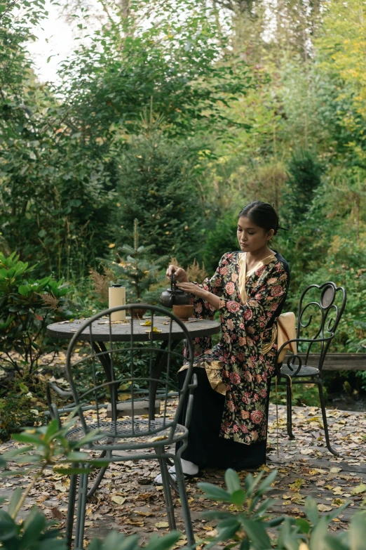 a woman sitting at a table in a garden, freida pinto, swirling gardens, holding a candle holder, cottagecore