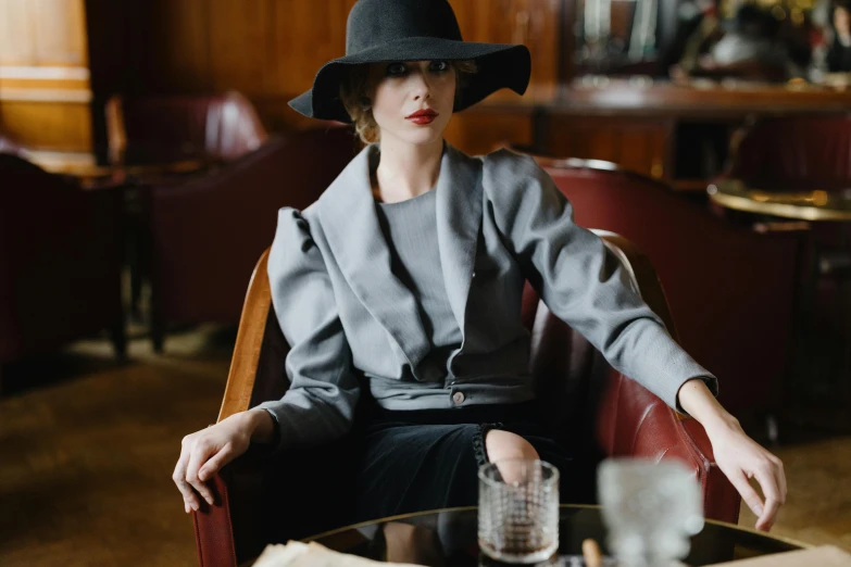 a woman sitting in a chair with a hat on, by Emma Andijewska, unsplash, renaissance, wearing black grey suit, sitting at the bar, voluminous sleeves, curated collection