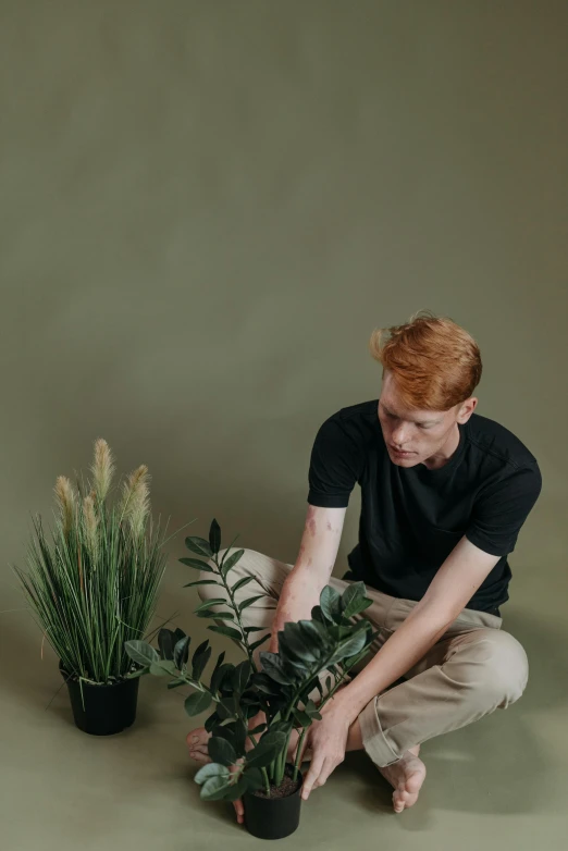 a man kneeling down next to a potted plant, trending on pexels, ginger hair, scattered props, plain background, muted green