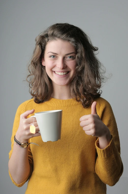 a woman in a yellow sweater holding a coffee cup, giving a thumbs up, on a gray background, uploaded, h3h3