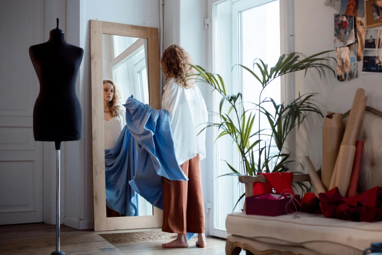 a woman standing in front of a mirror next to a mannequin, pexels contest winner, happening, blue cape, leaving a room, foliage clothing, inspect in inventory image