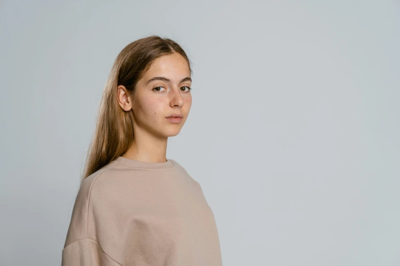 a woman standing in front of a gray background, a character portrait, by Attila Meszlenyi, trending on pexels, brown sweater, 1 6 years old, humanoid woman, light skin tone