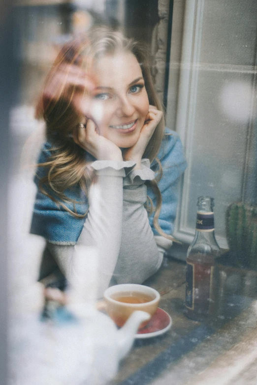 a woman sitting at a table with a cup of tea, a picture, by Adam Marczyński, pexels contest winner, looking through a window frame, smiling young woman, wearing turtleneck, hr ginger