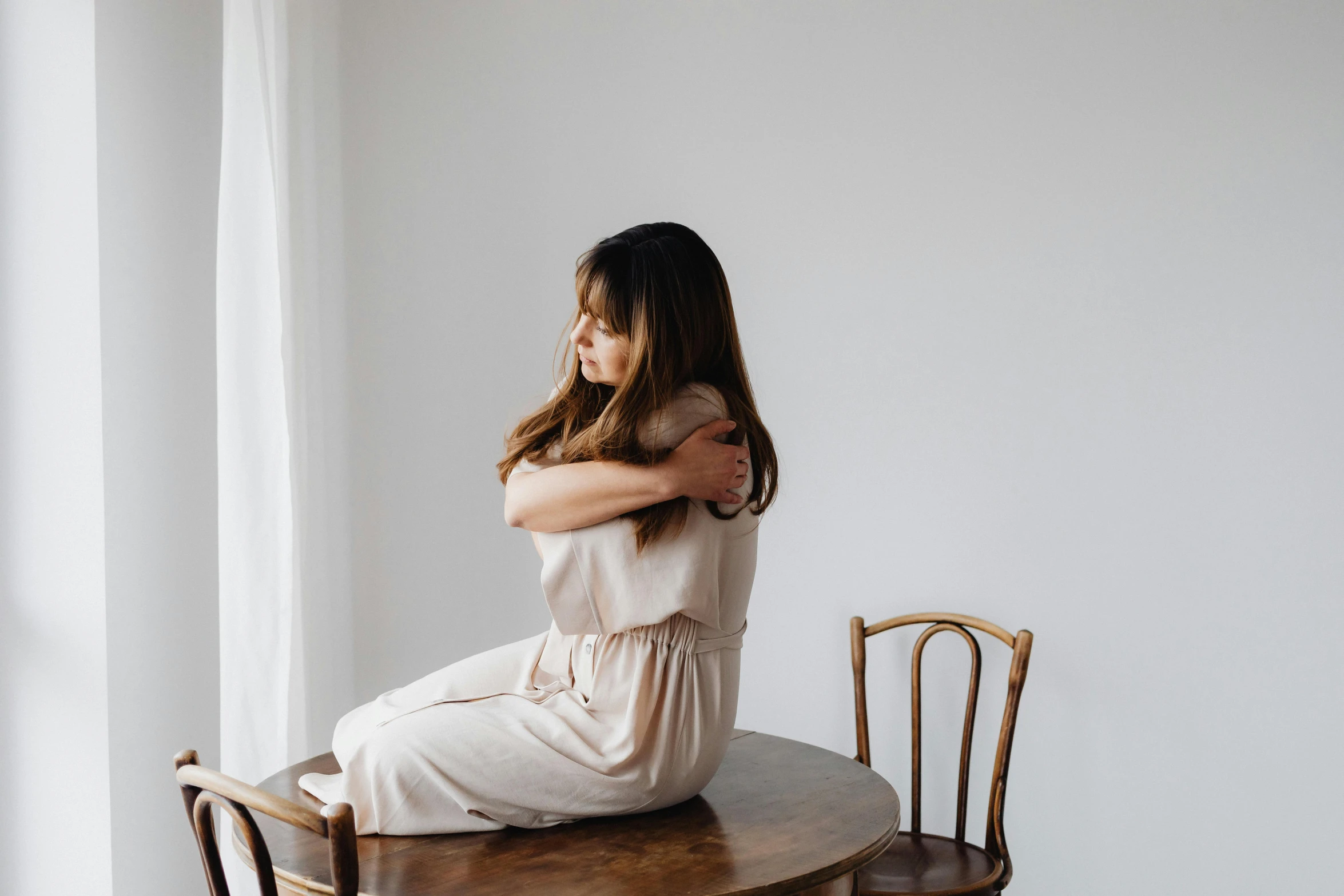 a woman sitting on top of a wooden chair, trending on pexels, romanticism, hugging, sitting on a mocha-colored table, in white room, waist reaching ponytail