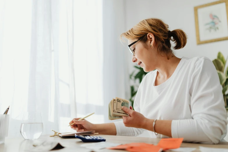 a woman sitting at a table with money and a calculator, a screenshot, pexels contest winner, wearing a white button up shirt, thumbnail, 🦩🪐🐞👩🏻🦳, mid - action