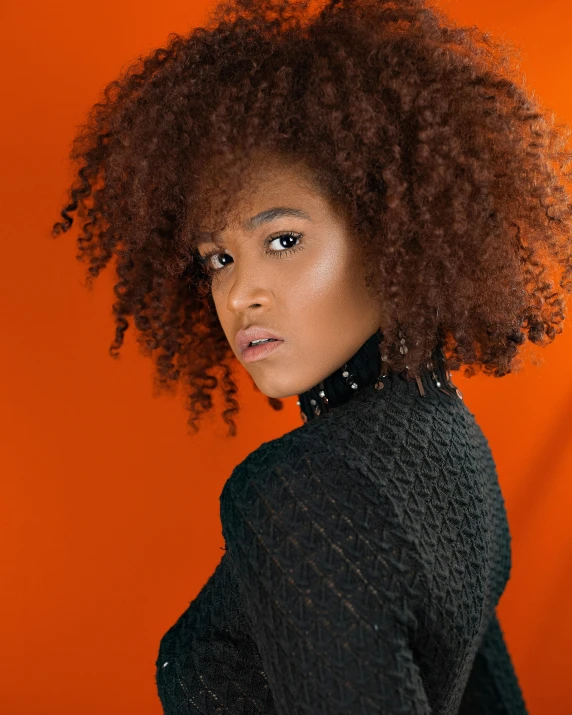 a woman with an afro standing in front of an orange background, trending on pexels, she has long orange brown hair, on black background, iridescent skin, looking back at the camera