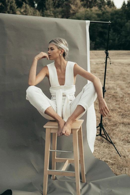 a woman sitting on a stool in a field, by Lucette Barker, trending on pexels, minimalism, white suit, short platinum hair tomboy, jumpsuit, magazine shoot