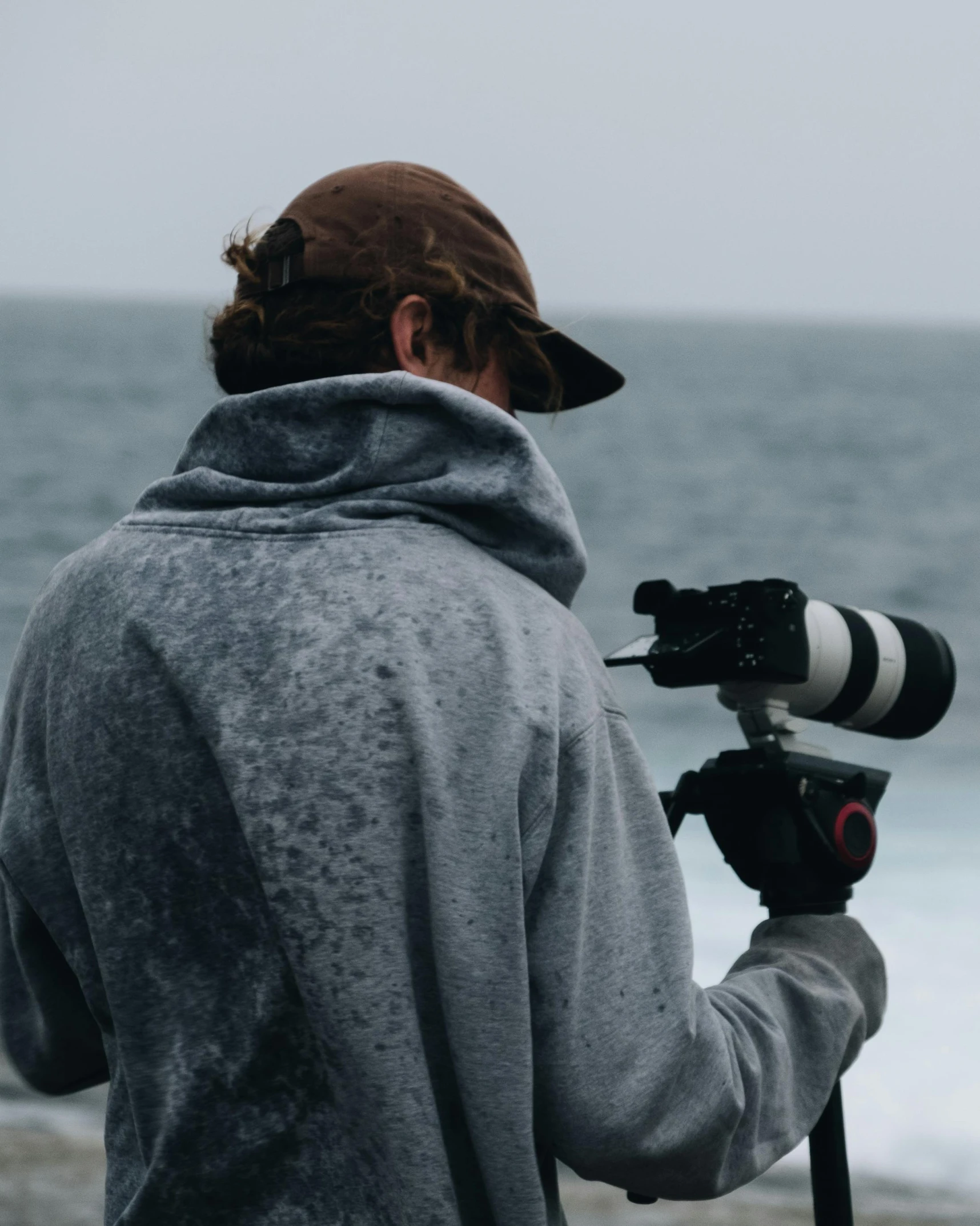 a man standing on top of a beach holding a camera, wearing a grey robe, water droplets on lens, he also wears a grey beanie, unsplash transparent