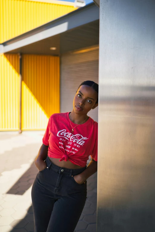 a woman leaning against a wall in front of a building, inspired by Dorothy Coke, trending on unsplash, red t-shirt, black teenage girl, soda, modelling