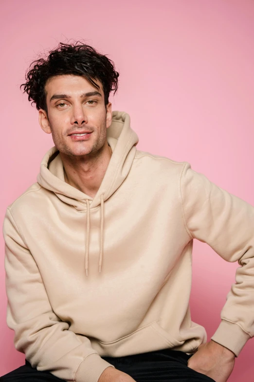 a man in a hoodie sitting on a stool, inspired by John Luke, trending on pexels, beige hoodie, wearing a pastel pink hoodie, curls on top, well-groomed model