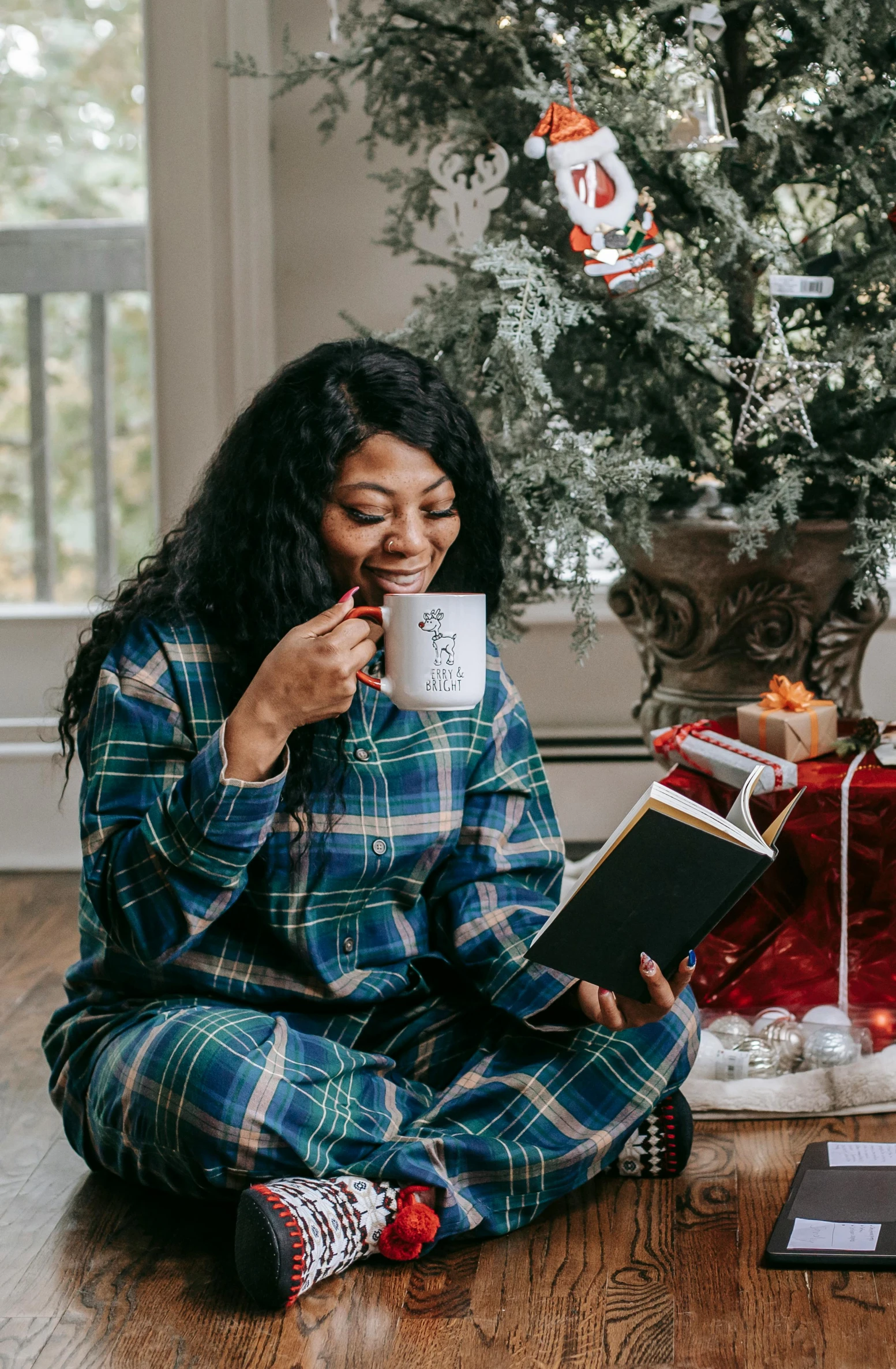 a woman sitting on the floor reading a book and drinking a cup of coffee, wearing festive clothing, flannel, riyahd cassiem, profile image