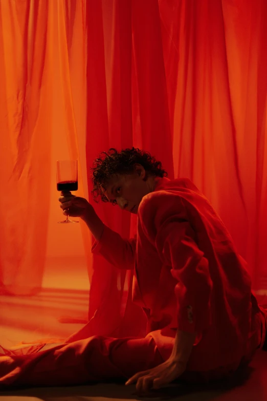 a woman sitting on the floor holding a glass of wine, an album cover, inspired by Nan Goldin, pexels contest winner, romanticism, red cloth around his shoulders, timothee chalamet, shades of aerochrome gold, ( ( theatrical ) )