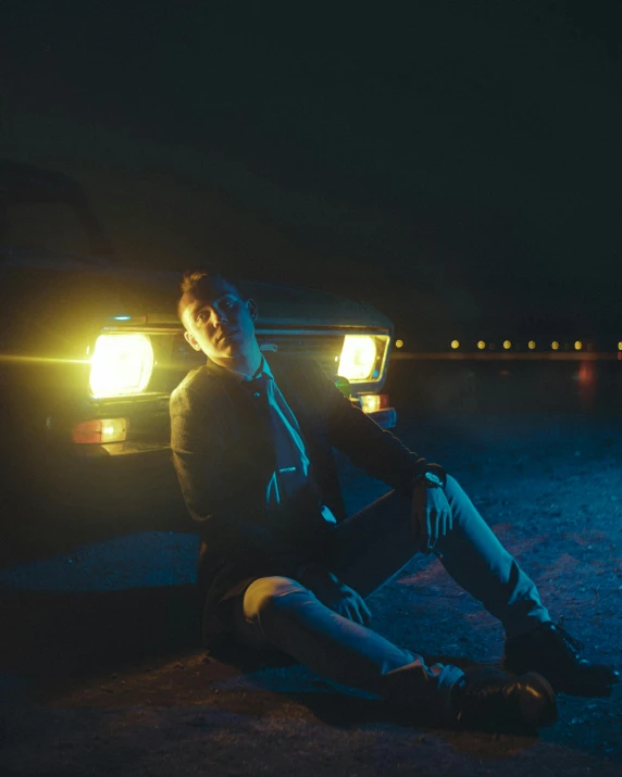 a man sitting on the ground next to a car, an album cover, by Elsa Bleda, unsplash, realism, bisexual lighting, face is brightly lit, julian ope, still from a music video