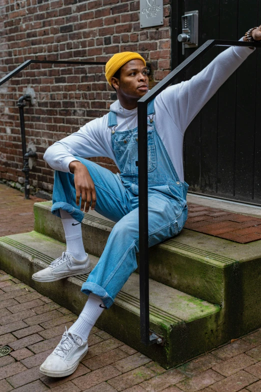 a man sitting on the steps of a building, by Jacob Toorenvliet, trending on pexels, realism, wearing blue jean overalls, nonbinary model, riyahd cassiem, yellow overall