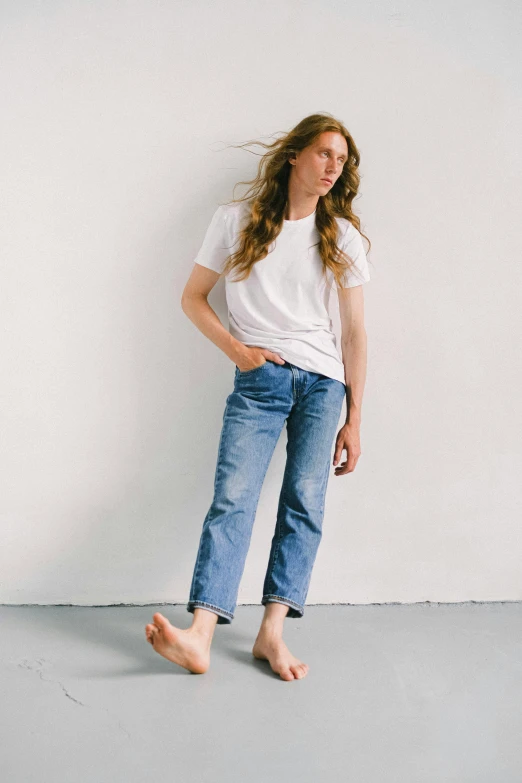 a woman standing in front of a white wall, unsplash, jeans and t shirt, androgynous male, sitting down casually, chillwave