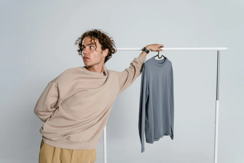 a man standing in front of a clothes rack, inspired by Viktor de Jeney, blue gray, wearing sweatshirt, posing like a falling model, taupe