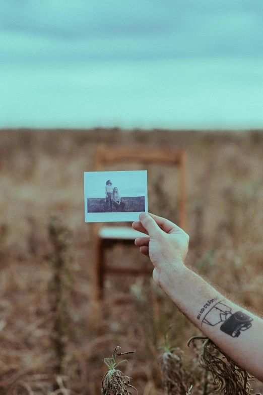 a man holding up an old photo in a field, a polaroid photo, pexels contest winner, surrealism, temporary tattoo, sitting on a chair, minimalist photo, with tattoos