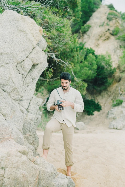 a man standing next to a large rock on a beach, holding a camera, wearing a linen shirt, instagram model, drew hill