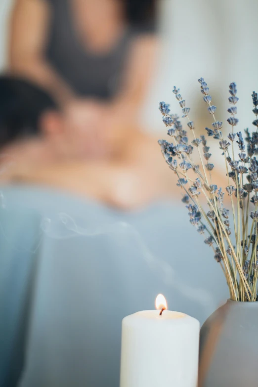 a candle sitting on top of a table next to a vase, lavender flowers, wearing a grey robe, embracing, candles in foreground